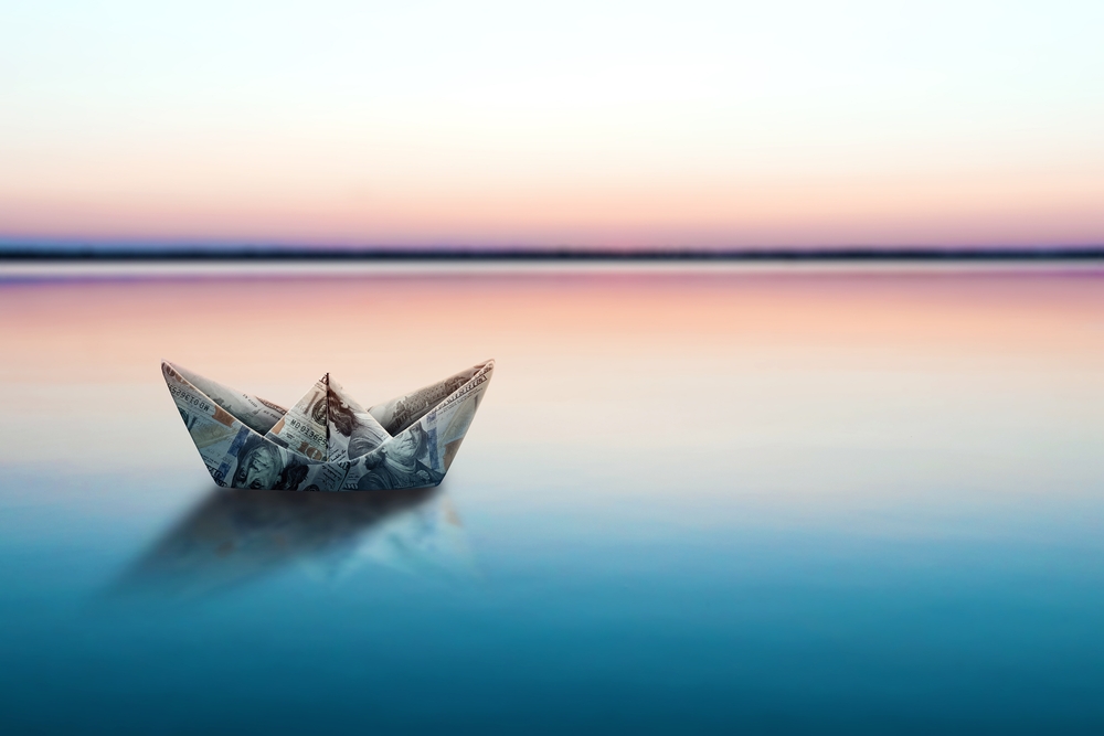 A paper boat made out of money sailing on water.